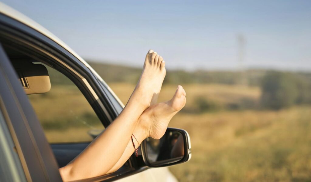 Barefoot feet resting outside a car window during a drive through a scenic countryside, symbolizing relaxed road trip vibes and comfort.
