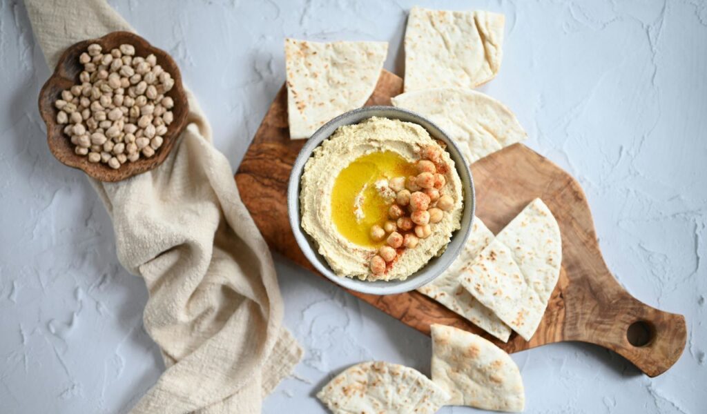 A bowl of hummus topped with chickpeas and olive oil is served on a wooden board with pieces of Druze pita bread and a bowl of dried chickpeas.