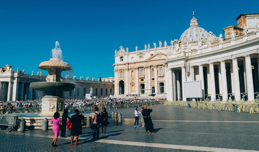  Visitors gathering at St. Peter's Square in Vatican City with a central fountain, the grand façade of St. Peter's Basilica, and a clear blue sky