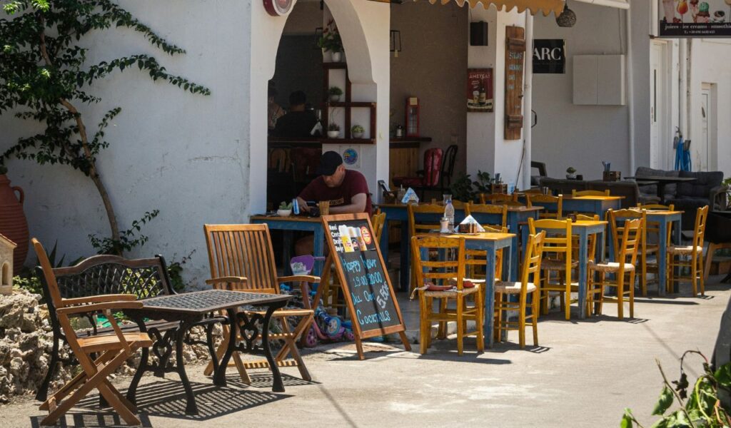 Outdoor Greek taverna with colorful wooden chairs and handwritten menu board, where a man sits enjoying a quiet local meal.