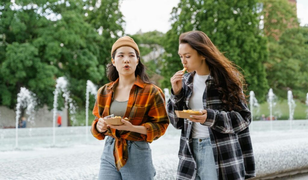 Two young women walk beside a fountain while eating from takeout containers. One is talking, and the other is eating a fry. Trees fill the background.