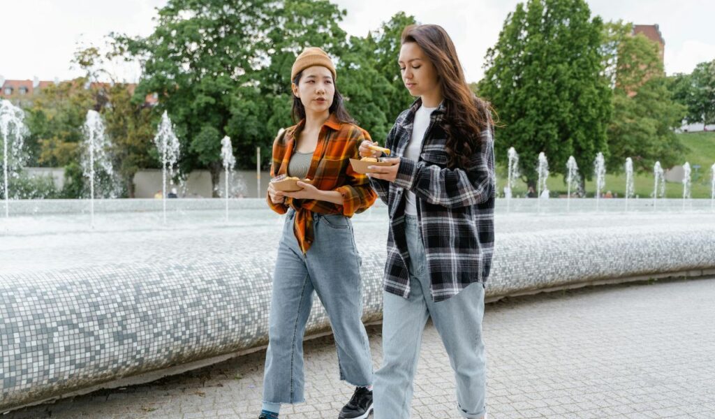 Two young women walk near a fountain while eating from takeaway containers, illustrating the concept of eating while walking in a public outdoor setting.