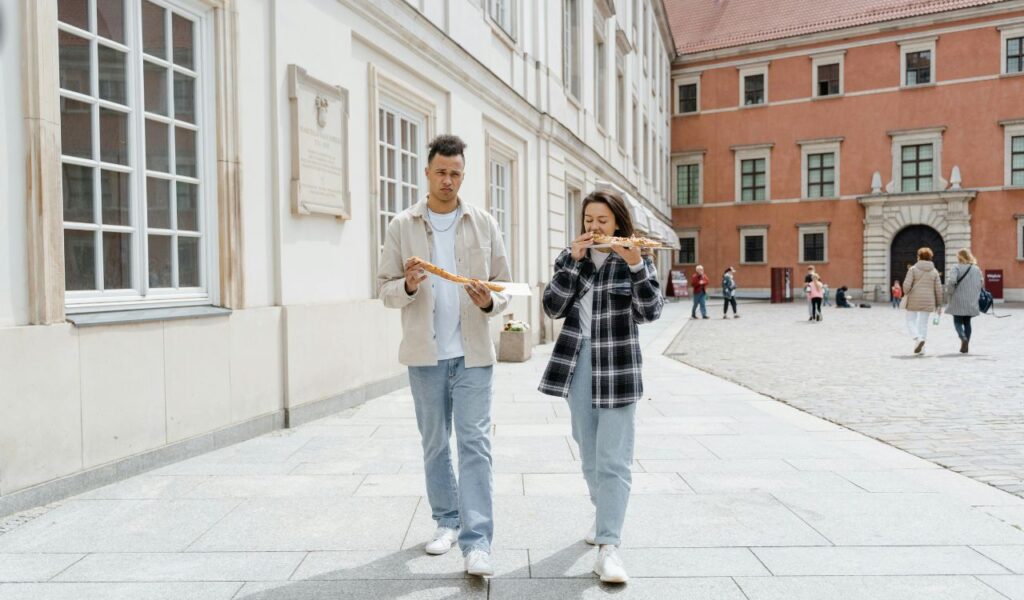 Two people walking outdoors while eating pizza slices, with historic buildings and other pedestrians in the background.