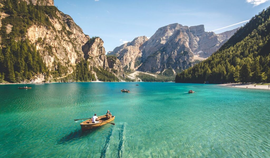 People rowing wooden boats on a clear turquoise lake surrounded by pine forests and rocky mountains under a bright blue sky.