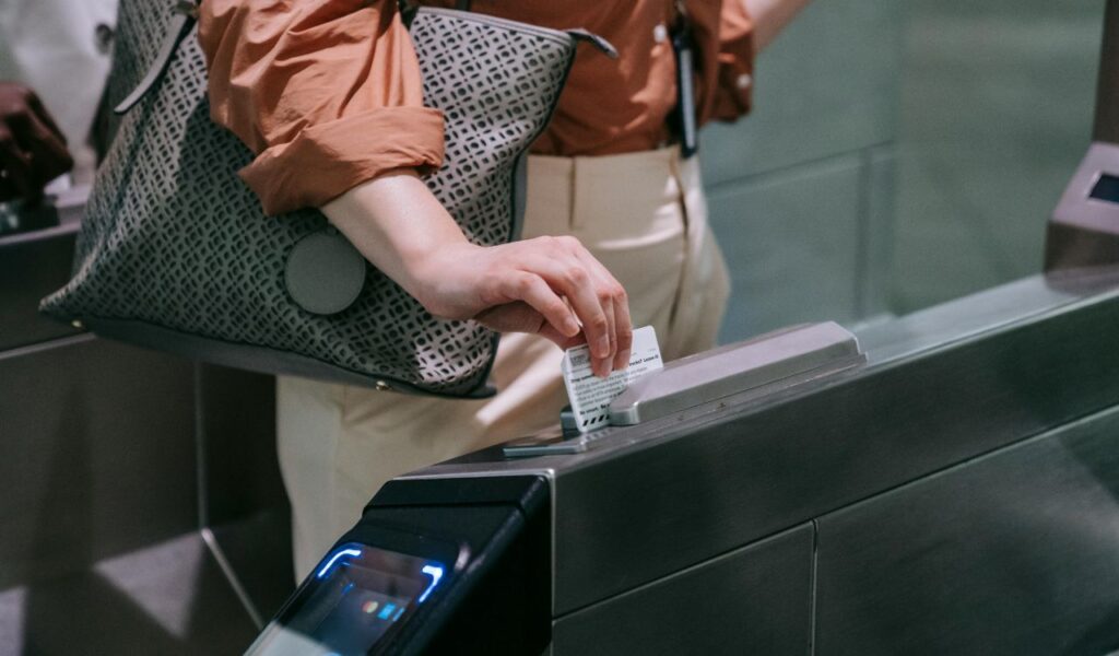 A person inserts a paper ticket into a turnstile reader at a transit station while carrying a patterned shoulder bag.