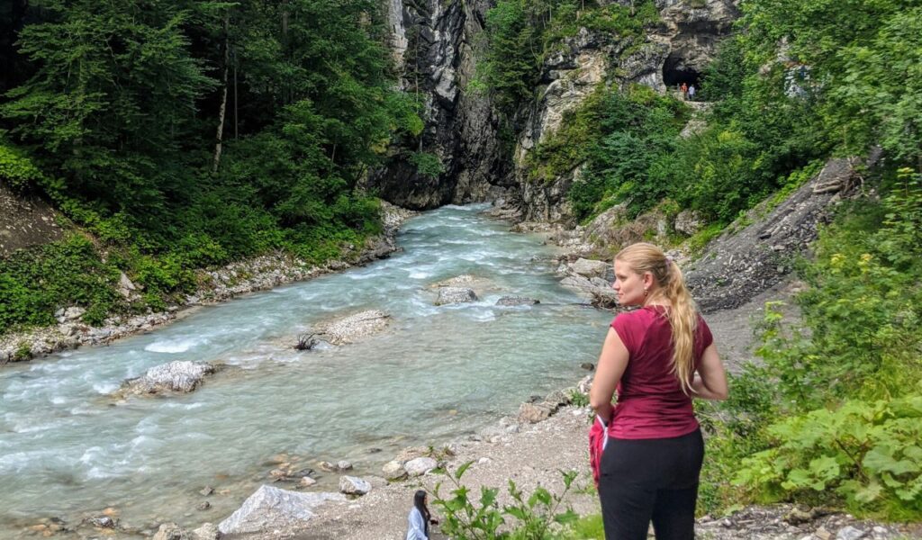 A woman standing near the turquoise river at Partnach Gorge in Bavaria, surrounded by lush green trees and rocky cliffs, with a cave entrance visible in the background.