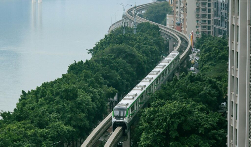 A modern passenger train travels along elevated tracks surrounded by lush green trees and urban buildings beside a calm river, showcasing scenic rail travel through a cityscape.