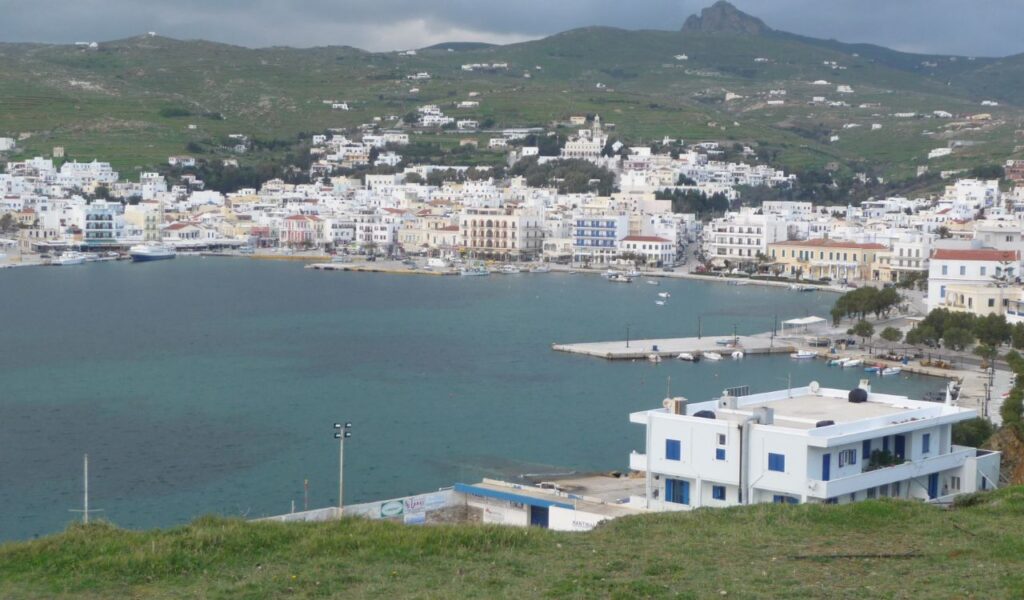 Panoramic view of Tinos island town with white buildings clustered around a peaceful harbor, backed by green hills and cloudy skies