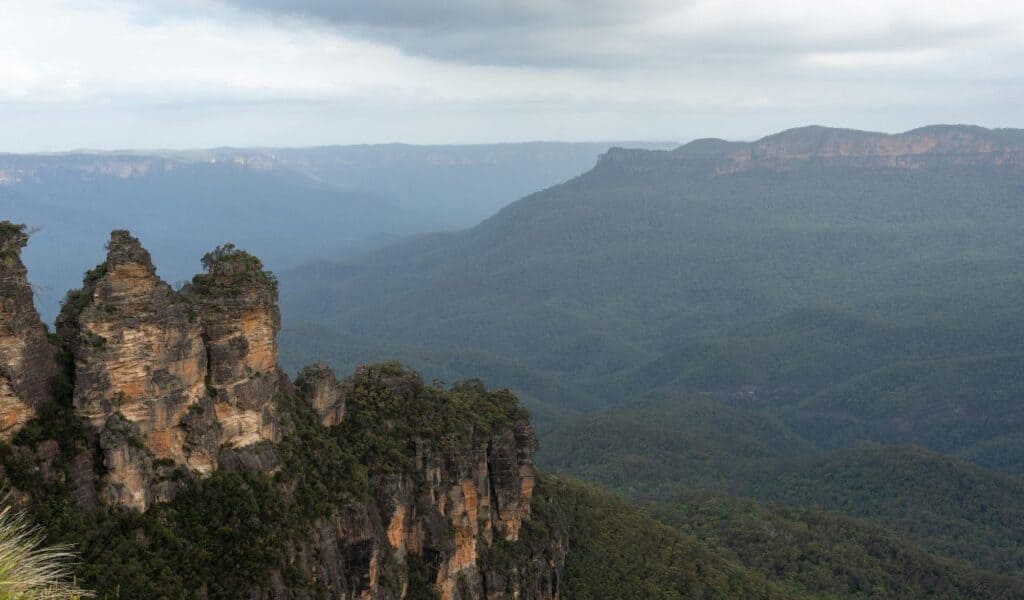 Dramatic rock formations rise from a densely forested mountain range under a cloudy sky, with layers of misty hills fading into the distance.