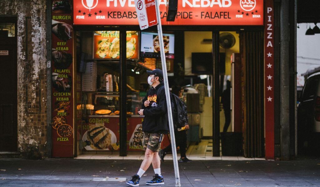 A person in a mask and hoodie walks past Five Star Kebab, a shop with red signage offering kebabs, pizza, pide, falafel rolls, and gozleme.