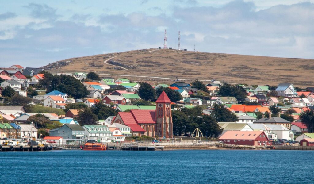 olorful houses and a red-roofed church in a coastal town on the Falkland Islands with rolling hills and radio towers in the background