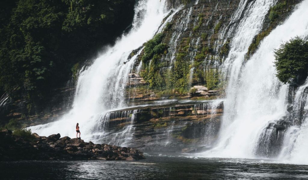 Woman stands on rocks at the base of a powerful, multi-tiered waterfall cascading down a lush, green cliff into a calm river.