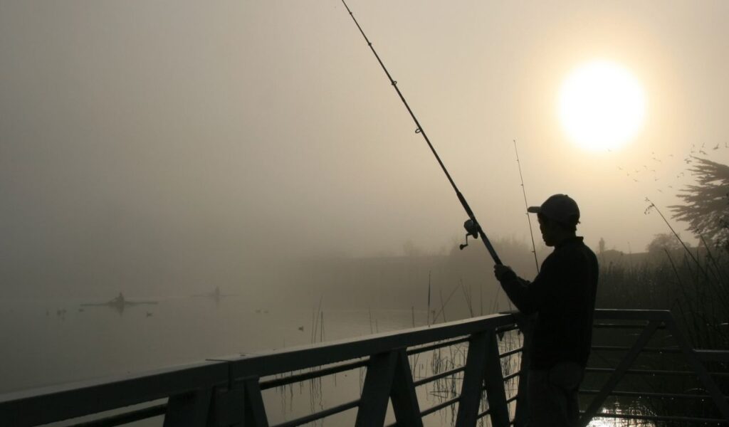 A person fishing off a bridge at sunrise, silhouetted against the foggy morning light with calm water and reeds in the background.
