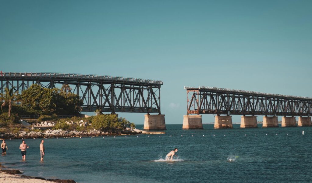 People swim and relax near a broken section of the historic Bahia Honda Rail Bridge in Florida, under a clear blue sky.
