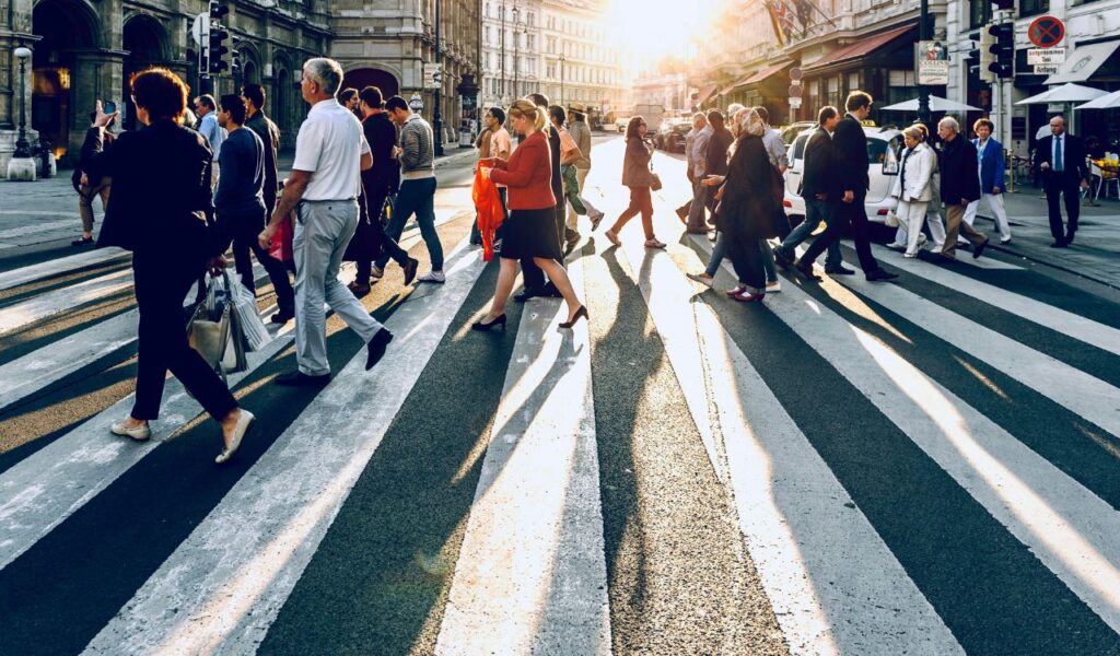A busy crosswalk filled with people walking in both directions during golden hour in a European city, casting long shadows on the street.