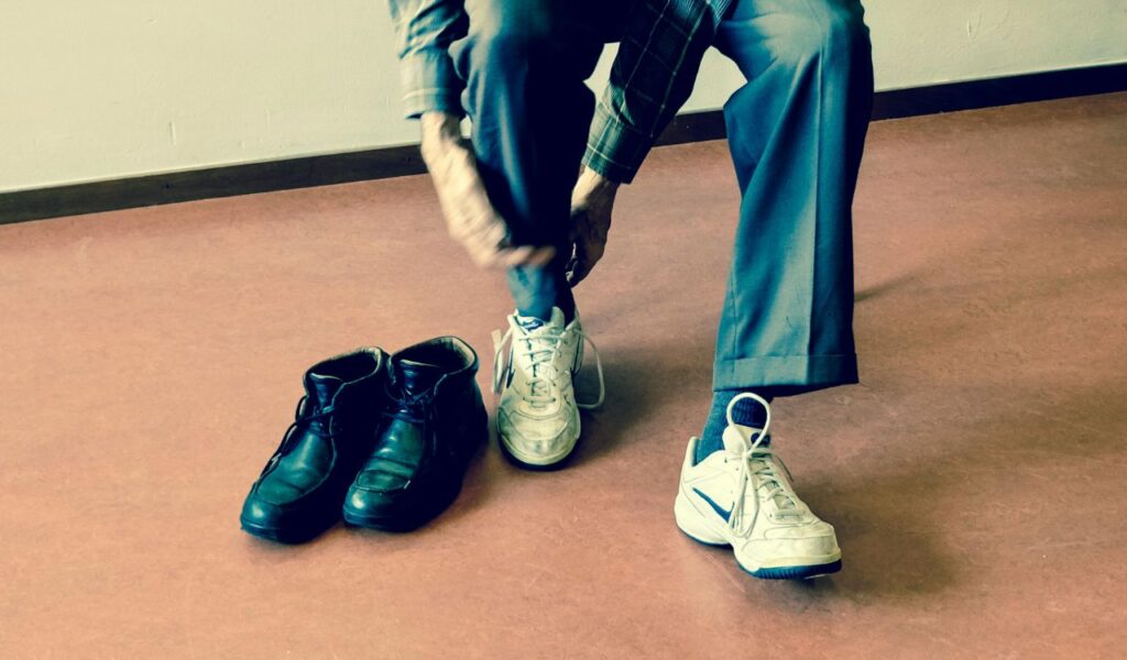 A person sitting indoors removes their sneakers, with a pair of black shoes placed on the floor nearby, representing the practice of taking off shoes before entering homes or indoor spaces.