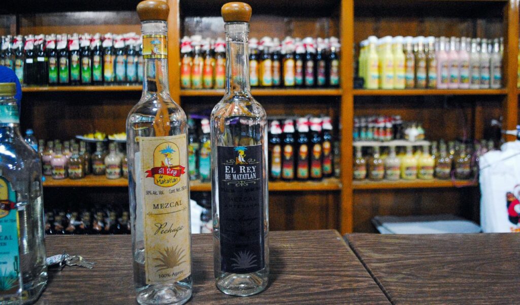 Two bottles of mezcal sit on a wooden bar counter, with shelves of colorful liquor bottles lined up in the background.