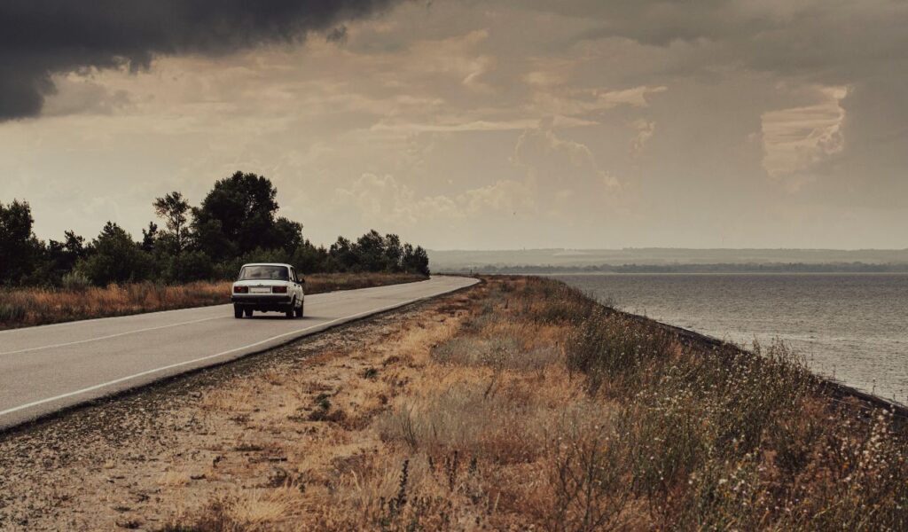 A lone car drives along an empty rural road beside a large body of water under a dramatic, cloudy sky.