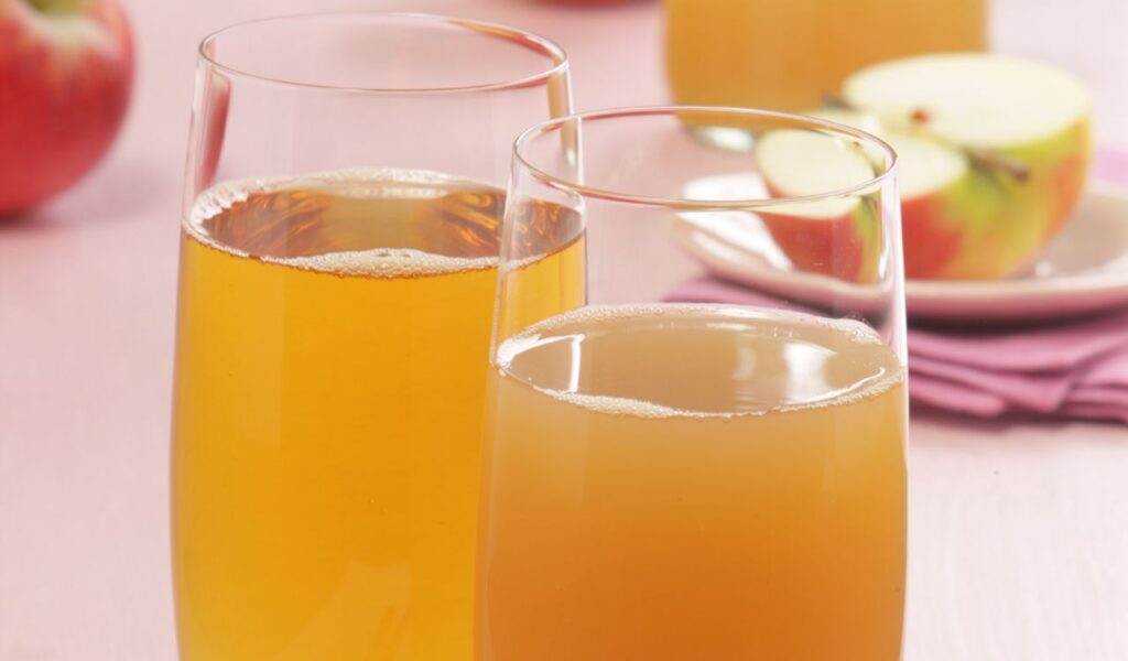Two glasses of fruit juice, with a close-up view of the golden liquid and apple slices in the background