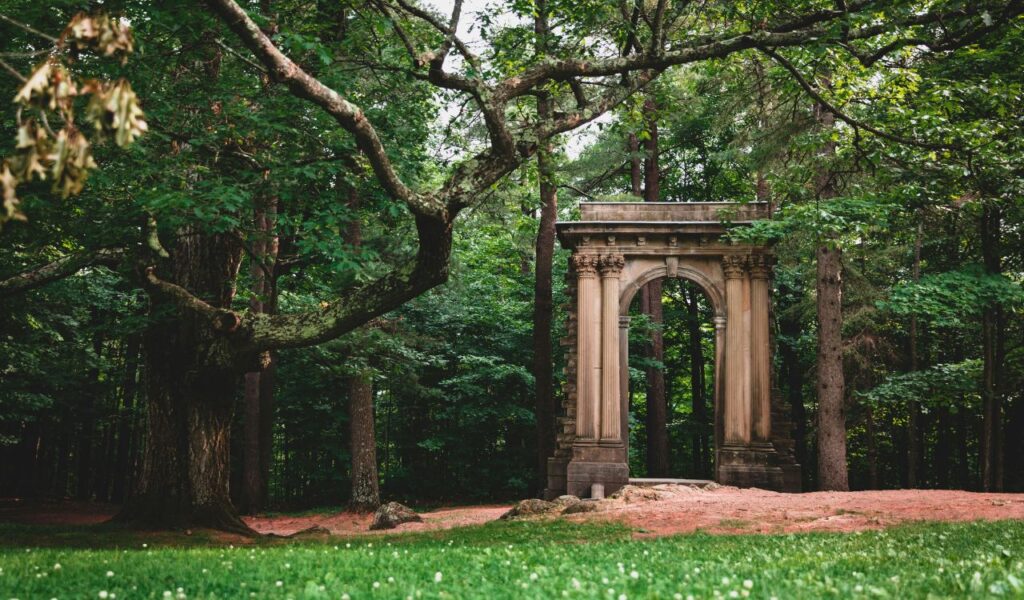 A lone stone arch stands among tall trees in Gatineau Park, surrounded by grass, dirt paths, and thick forest