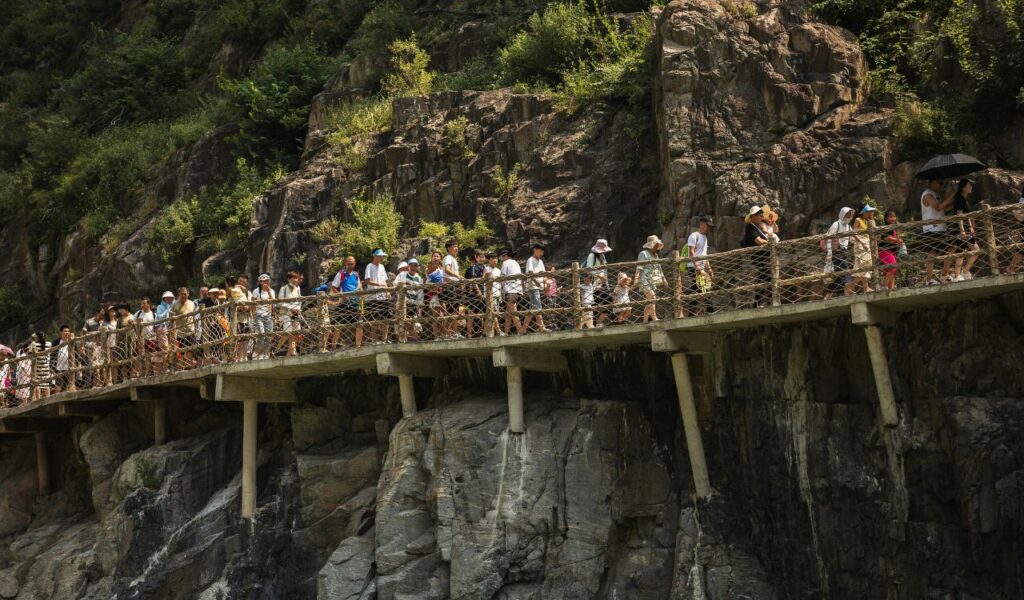 Crowds of hikers walking along a narrow elevated pathway with railings built into a rocky cliffside at Partnach Gorge, surrounded by rugged terrain and greenery.
