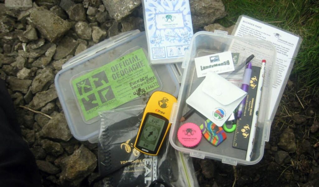 A geocaching kit spread out on rocky ground, including a GPS device, logbooks, small trinkets, and official geocache containers.