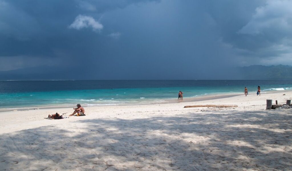 Tourists relax and stroll along the white sandy beach of Gili Meno, Indonesia, with turquoise water and stormy skies.