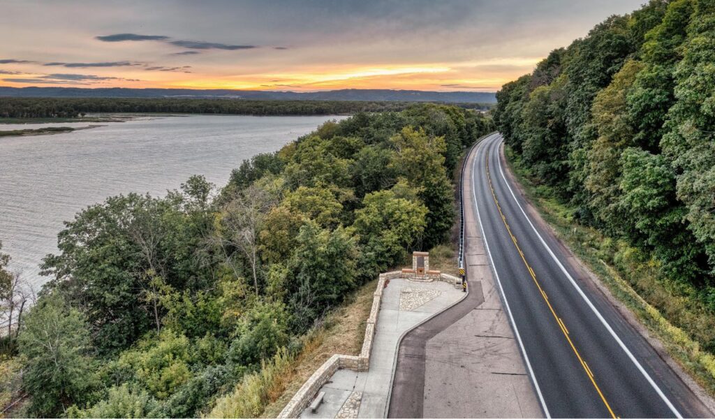 Scenic view of the Great River Road running alongside the Mississippi River, bordered by lush green forests under a colorful sunset sky.