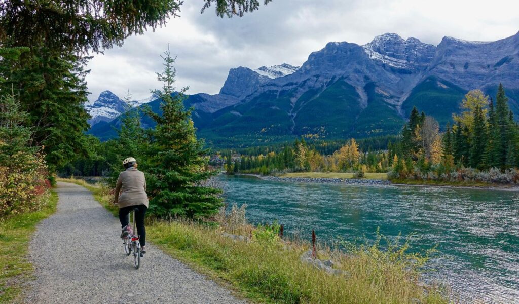 A cyclist rides along the Greenbrier River Trail beside a calm river, with pine trees and rugged mountain peaks in the background.