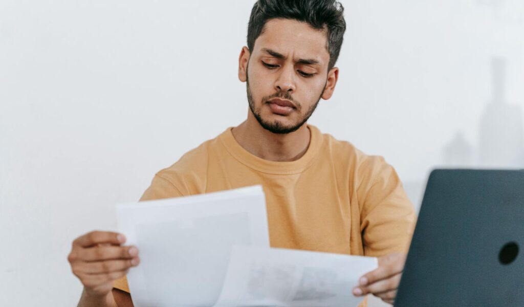 A man in a beige shirt sits at a desk, seriously reviewing printed documents next to an open laptop, suggesting offline decision-making.