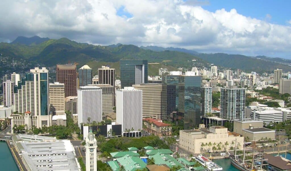 Honolulu city skyline with modern high-rises backed by lush green mountains and partly cloudy skies in Hawaii.