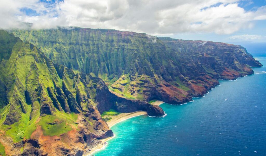 Aerial view of Kauai’s dramatic Na Pali Coast with lush green cliffs, turquoise water, and a hidden beach below.