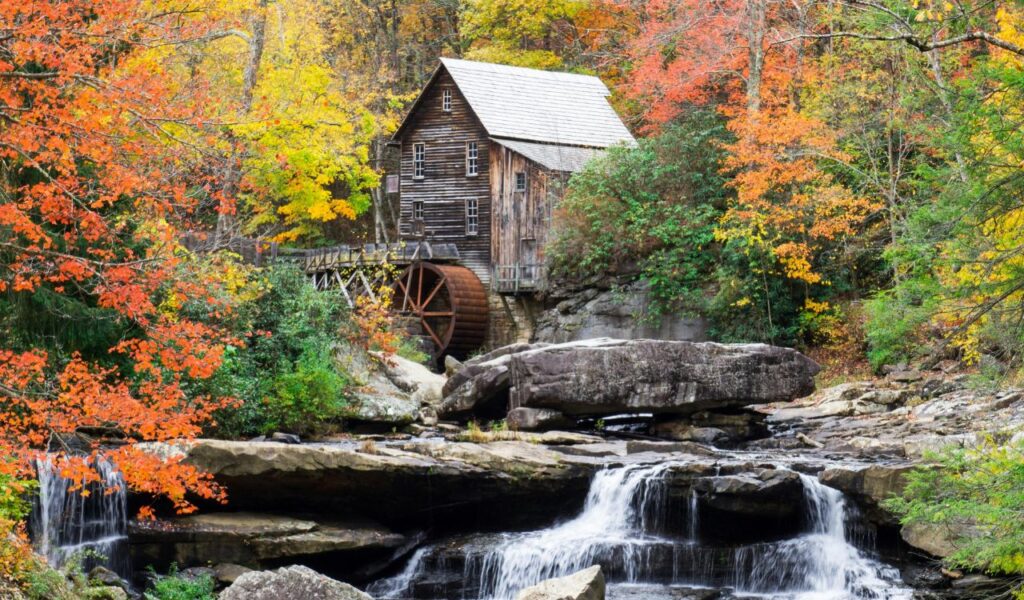 A historic wooden grist mill with a large waterwheel sits beside a cascading stream, surrounded by vibrant autumn foliage in Hawks Nest State Park, Ansted.
