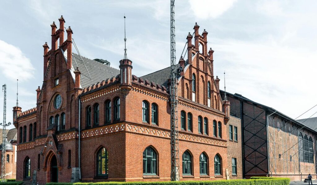 A large red-brick industrial building with Gothic architectural elements, tall chimneys, arched windows, and a slate roof, set against a partly cloudy sky.