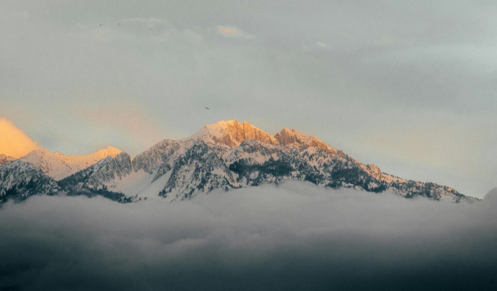 Snow-covered peaks of the Henry Mountains rise above thick clouds, glowing softly in the warm light of sunrise or sunset.