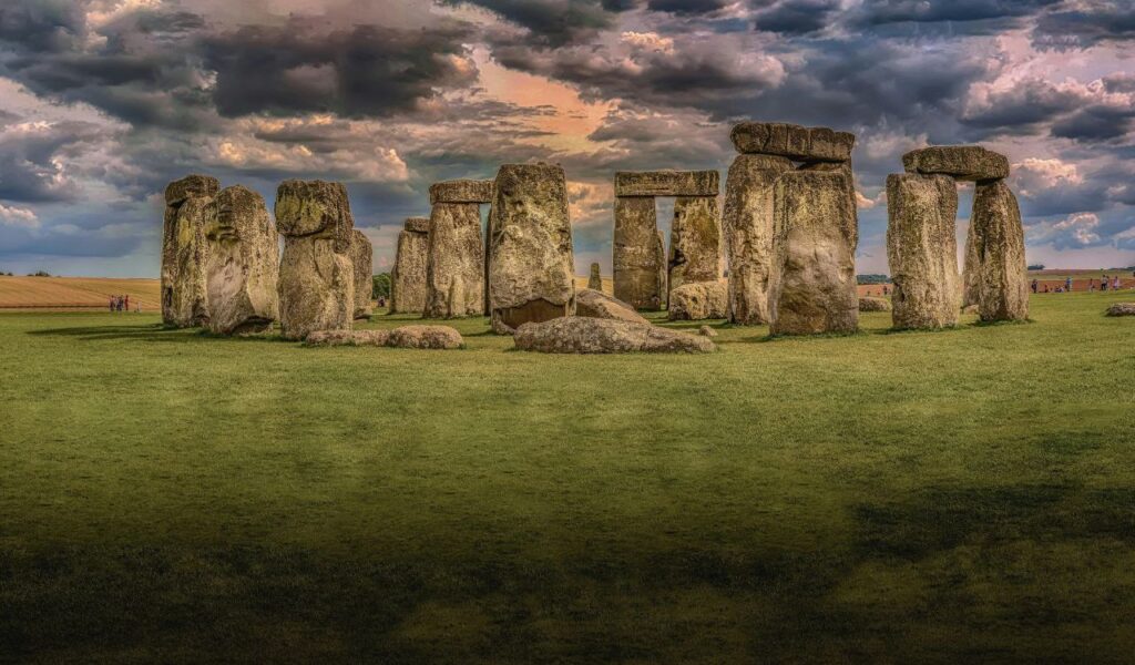 Ancient stone circle of Stonehenge under a dramatic cloudy sky at sunset, surrounded by open grassy fields and distant visitors.
