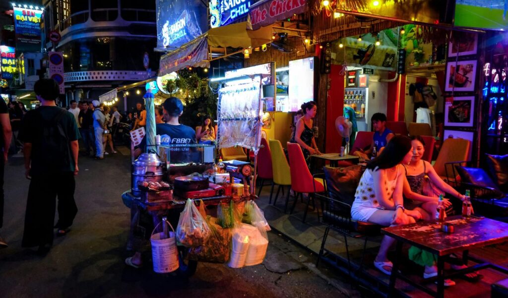 A lively night market scene in Ho Chi Minh City with street food stalls, neon lights, and people dining and walking along the bustling street
