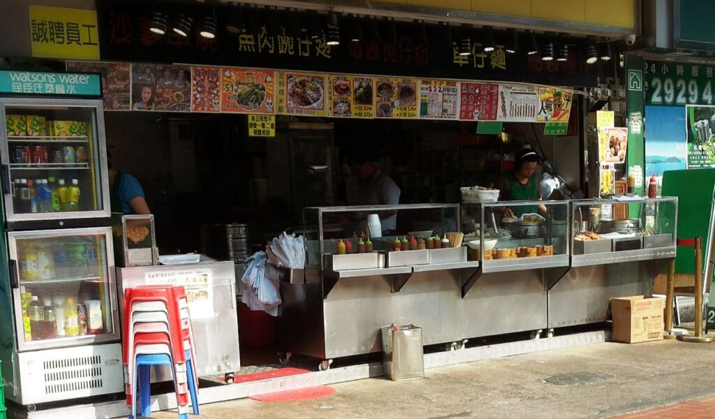 Outdoor Hong Kong street food stall with glass display cases, bottled drinks, sauces, and vendors preparing local dishes.