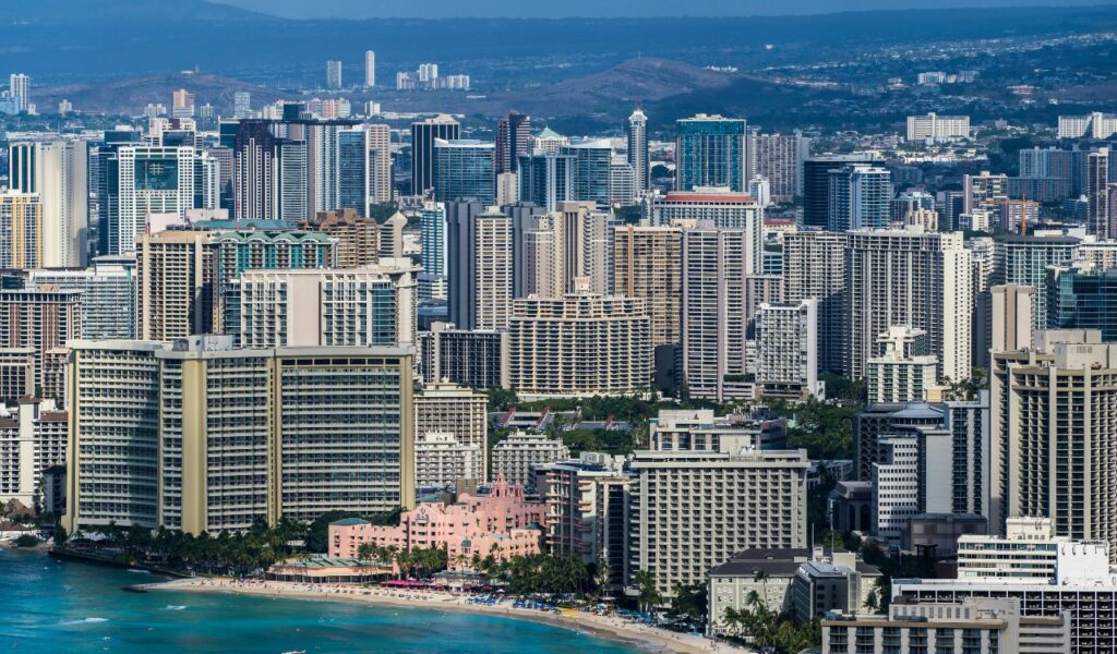 Aerial view of Honolulu, Hawaii, featuring high-rise buildings along Waikiki Beach with turquoise ocean waters and distant mountains.