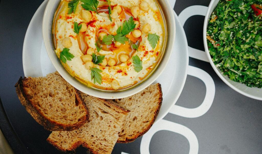 A bowl of hummus topped with chickpeas, parsley, and paprika sits on a plate with slices of rustic bread, next to a bowl of fresh green salad.