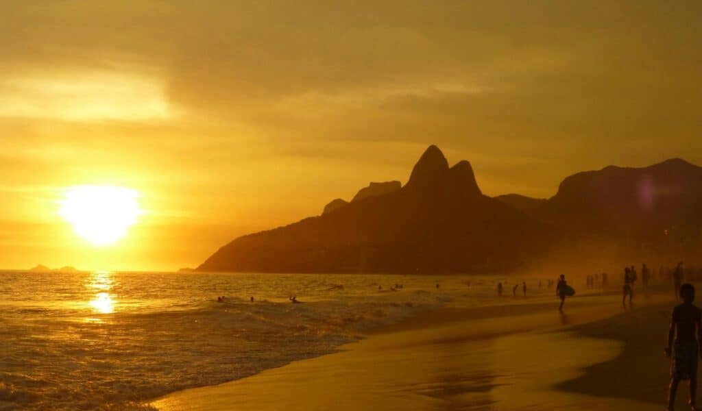 People walking and swimming along a beach at sunset with golden light reflecting on the sand and ocean, and dark mountain silhouettes in the background.