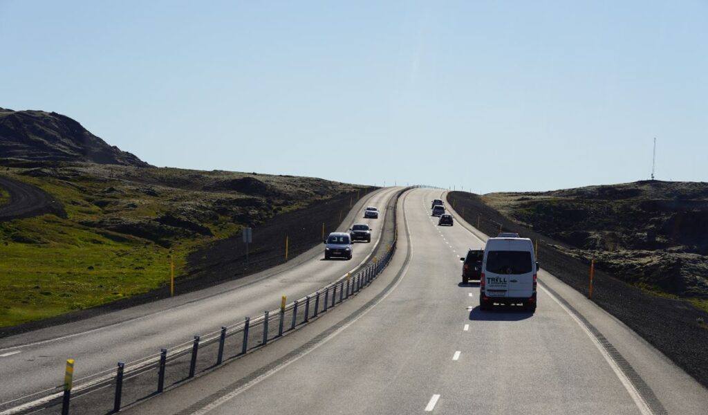 Vehicles travel along a smooth, two-lane highway winding through a rugged, hilly landscape under a clear blue sky.