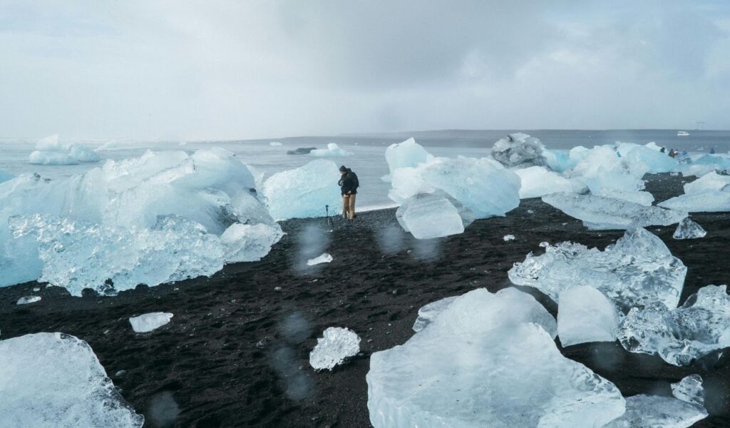 A person stands on a black sand beach surrounded by large, scattered icebergs under a cloudy sky in Iceland.