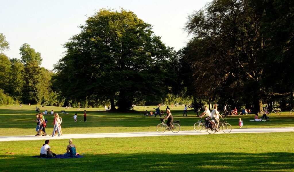 People enjoying a sunny day at Idlewild Park in Pennsylvania, with families picnicking, cyclists riding along a path, and lush green trees in the background.