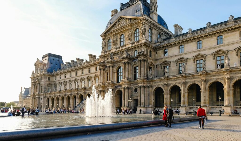Visitors gather around a large fountain in front of a grand historic building with arched windows and ornate sculptures at the Louvre Museum in Paris.
