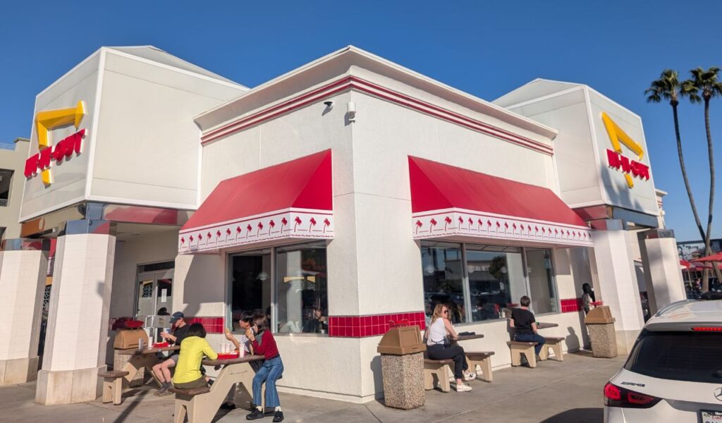 People dining outside an In-N-Out Burger restaurant with red awnings and palm tree design.