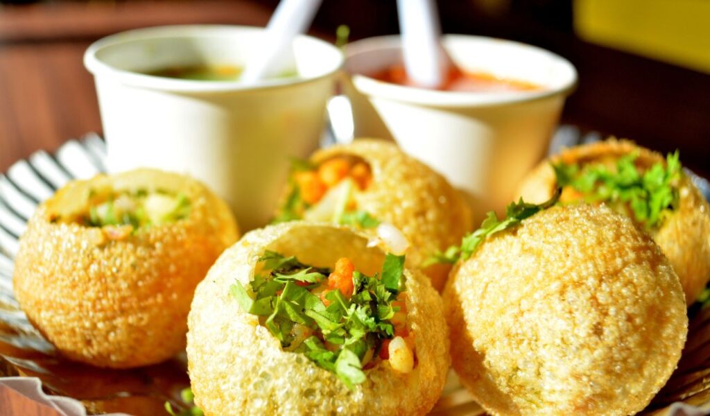 Close-up of Indian pani puri filled with spiced potatoes and herbs, served with tangy flavored water in white cups behind.