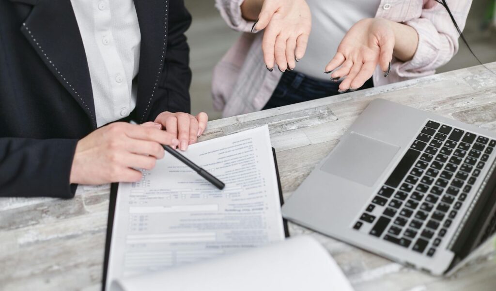 Two people review a form together at a desk with a laptop, one pointing with a pen while the other gestures in confusion.
