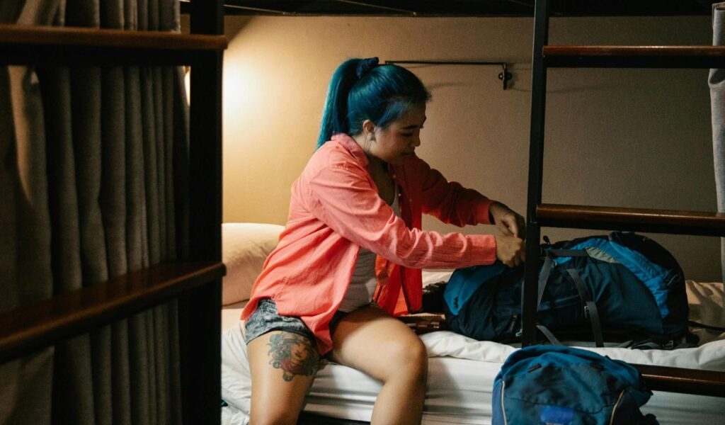 Backpacker sitting on a hostel bunk bed, unpacking a bag, preparing to settle in for the night.