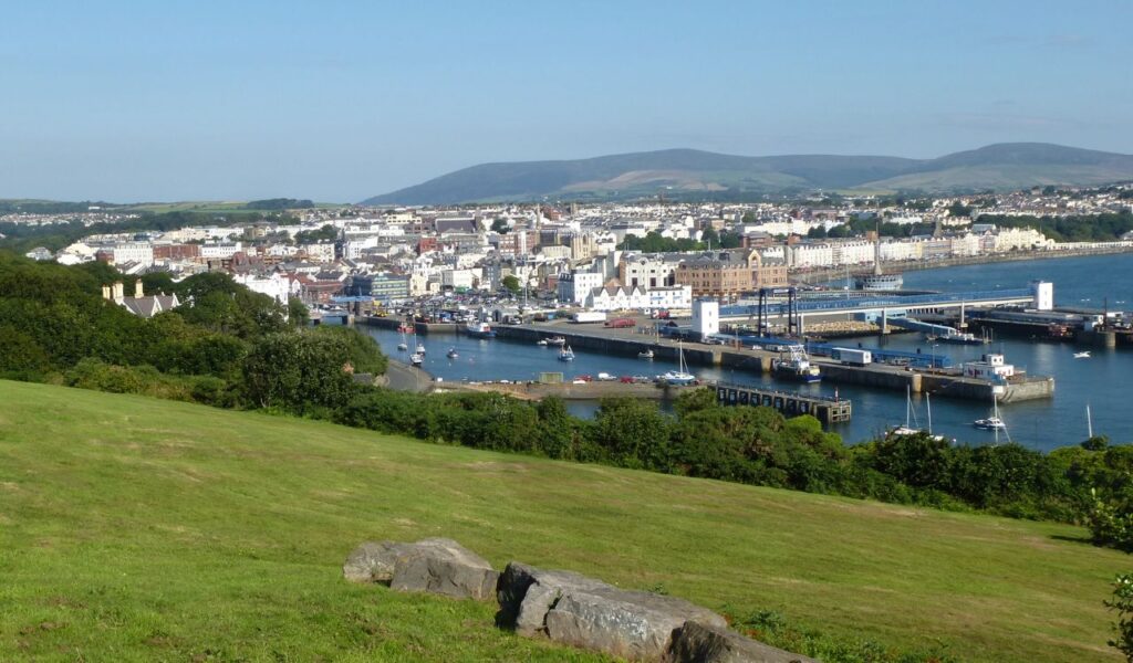 A scenic view of Douglas, the capital of the Isle of Man, with a harbor, moored boats, and buildings backed by rolling hills.